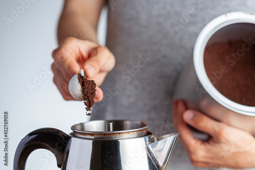 A caucasian woman is taking a teaspoon of ground coffee from a jar using a plastic measuring spoon and putting it into a percolator for brewing homemade morning coffee. Closeup selective focus image. photo