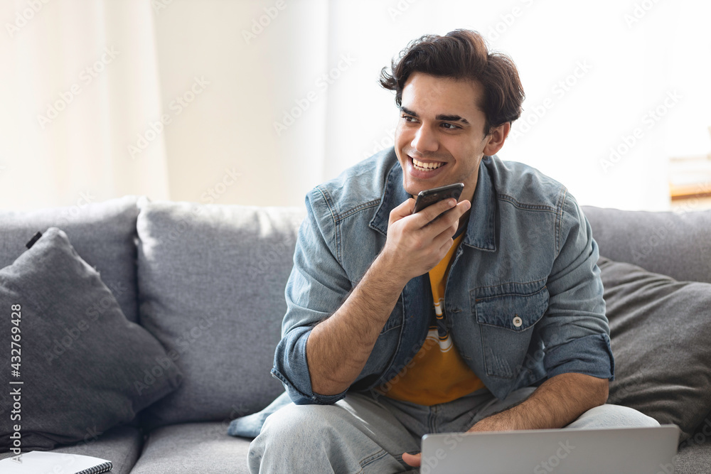 Young successful freelance entrepreneur working remotely at home using laptop and smartphone. Man talking on the phone while sitting on the couch at home