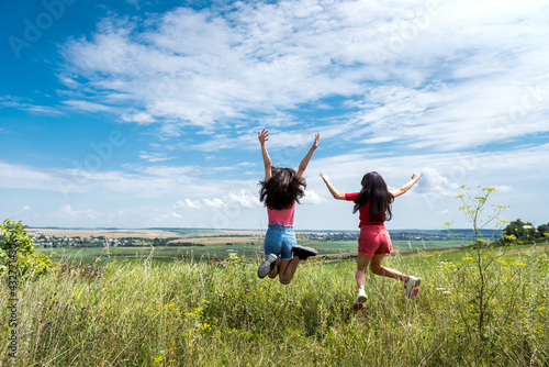 two happy slim smiling young women jumping together on rural field on the blue sky, summer time