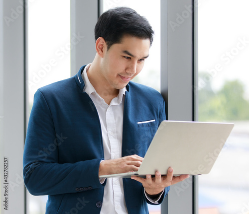 A portrait of a relaxed young businessman in a casual suit standing, holding a laptop and looking to screen smiling happily. Business success concept..