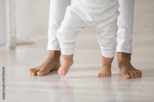 Crop close up of caring African American mom hold small toddler daughter hands make first steps together. Little baby kid child learn walking on wooden floor in home with mother. Childhood concept.