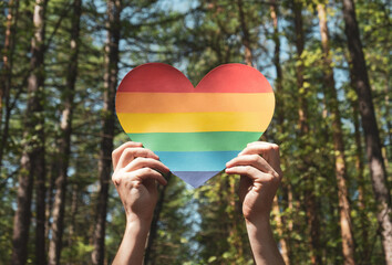 Man hands hold heart in colors of rainbow in the forest against the sky. LGBT rights, Pride month, love and unity with nature concept. Close up