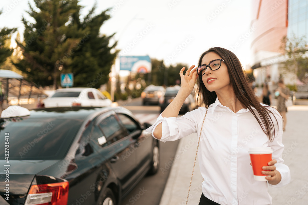 Attractive businesswoman watching to call a cab, confident trendy dressed female manager hailing on road catching taxi holding coffee to go getting to work