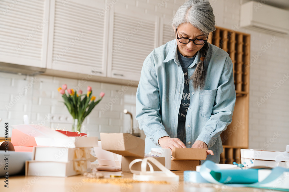 Mature grey woman smiling while wrapping presents in kitchen at home Mature grey woman smiling while wrapping presents in kitchen at home