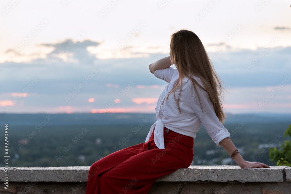 Young beautiful woman on the side view panoramic platform. Sunset and forest on the distance Young beautiful woman on the side view panoramic platform. Sunset and forest on the distance