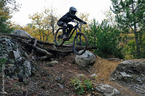 A female mountain biker at the wheel of his mountain bike makes a trick in jumping on the springboard of the downhill mountain path in the autumn forest.