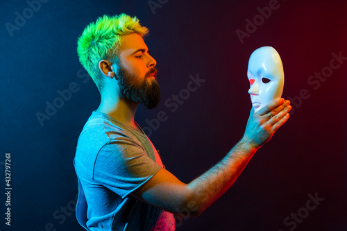 Side view of confident serious man in denim overalls, holding and looking at white mask in hands with attentive look, trying to understand hiding personality. Colorful neon light, indoor studio shot.