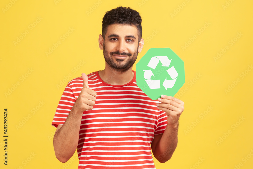 Positive smiling man with beard showing thumbs up gesture holding green waste recycling symbol, satisfied with environmental safety. Indoor studio shot isolated on yellow background Positive smiling man with beard showing thumbs up gesture holding green waste recycling symbol, satisfied with environmental safety. Indoor studio shot isolated on yellow background