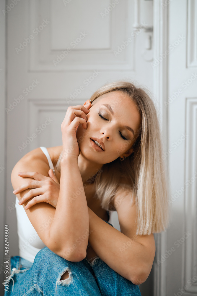 Beautiful woman posing in white body and blue jeans siting near white door at home Beautiful woman posing in white body and blue jeans siting near white door at home