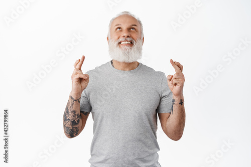 Smiling old man with tattoos looking up in sky, make wish and cross fingers for good luck, praying, begging god for fortune or dream come true, standing against white background
