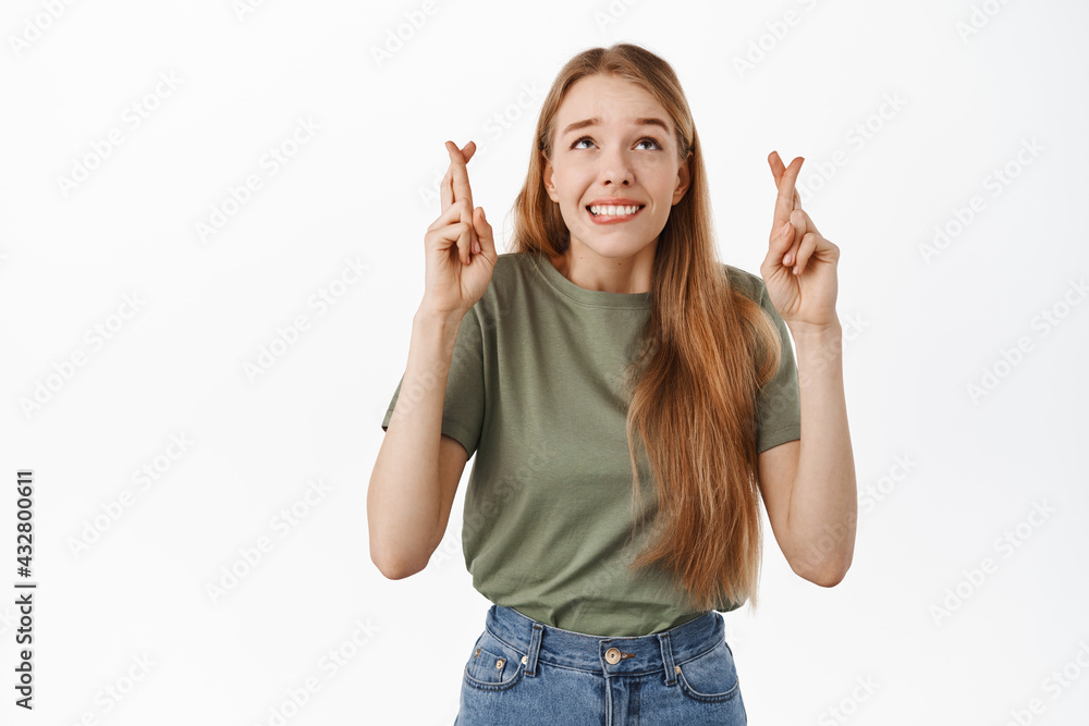 Nervous girl student praying with fingers crossed, looking worried up and pleading to pass exam, standing against white background Nervous girl student praying with fingers crossed, looking worried up and pleading to pass exam, standing against white background