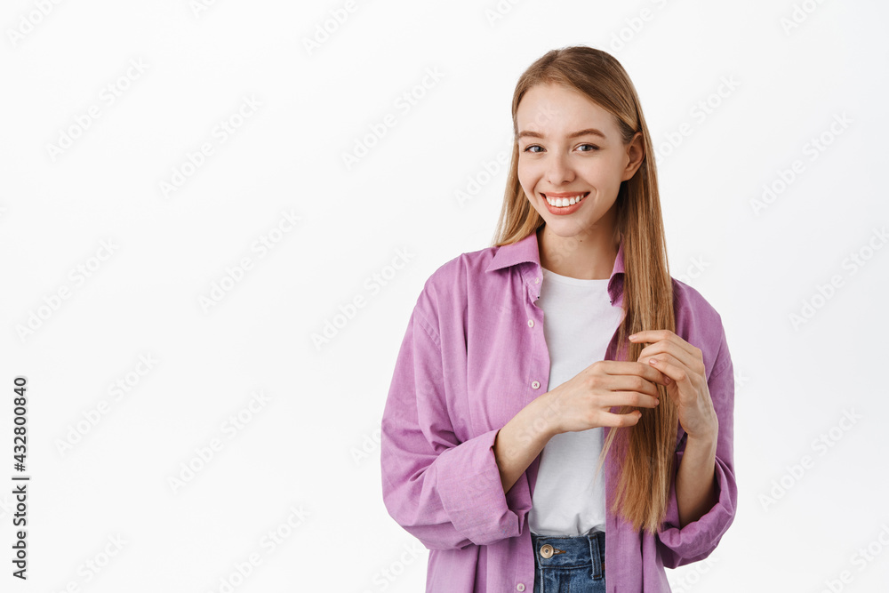 Happy cute blond woman touching her straight healthy hair, smiling white teeth and looking at camera, standing in trendy summer clothes against white background Happy cute blond woman touching her straight healthy hair, smiling white teeth and looking at camera, standing in trendy summer clothes against white background