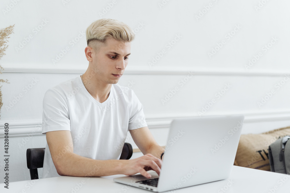 Photo of a blonde young man at the table at home talking using laptop computer. Photo of a blonde young man at the table at home talking using laptop computer.