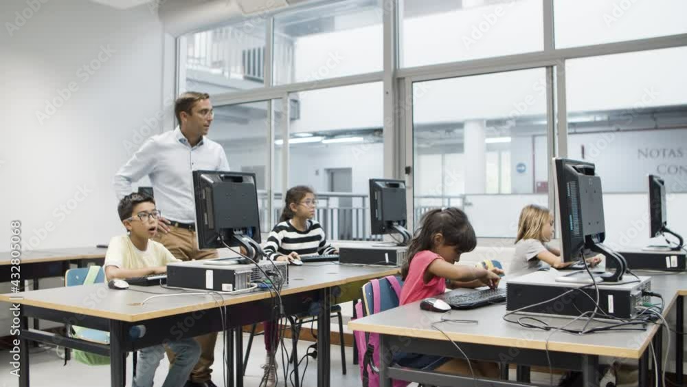 Computer science teacher watching as pupils doing task. Male teacher standing behind children in classroom. Multiethnic kids sitting, working on computer. Education, informatics concept. Computer science teacher watching as pupils doing task. Male teacher standing behind children in classroom. Multiethnic kids sitting, working on computer. Education, informatics concept.