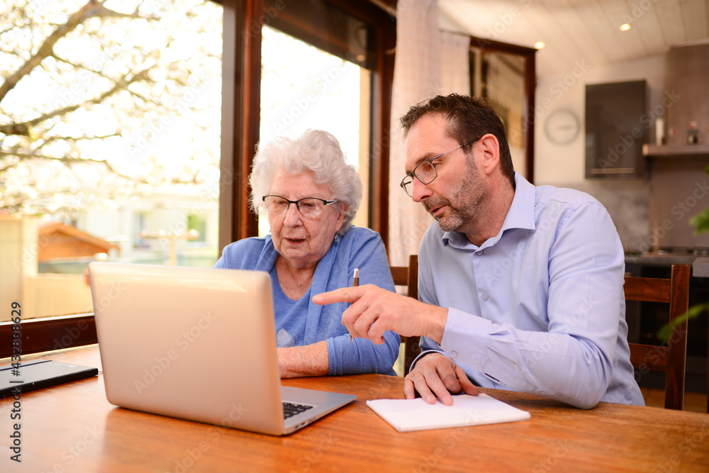 mature man helping elderly senior woman at home with paperwork and computer internet lesson - obrazy, fototapety, plakaty 