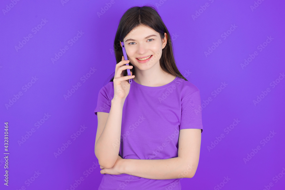 Portrait of a smiling young beautiful Caucasian woman wearing purple T-shirt over purple wall talking on mobile phone. Business, confidence and communication concept. Portrait of a smiling young beautiful Caucasian woman wearing purple T-shirt over purple wall talking on mobile phone. Business, confidence and communication concept.