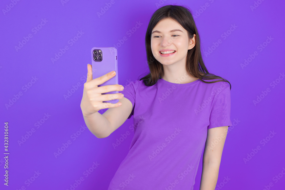 Isolated shot of pleased cheerful young beautiful Caucasian woman wearing purple T-shirt over purple wall, makes selfie with mobile phone. People, technology and leisure concept Isolated shot of pleased cheerful young beautiful Caucasian woman wearing purple T-shirt over purple wall, makes selfie with mobile phone. People, technology and leisure concept