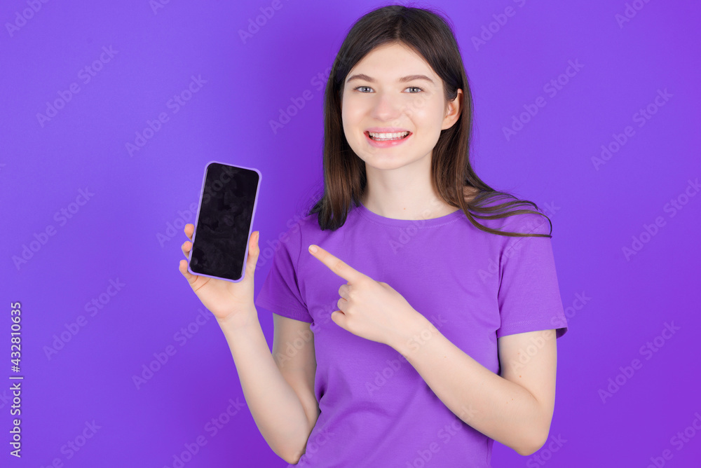 Smiling young beautiful Caucasian woman wearing purple T-shirt over purple wall showing and pointing at empty phone screen. Advertisement and communication concept. Smiling young beautiful Caucasian woman wearing purple T-shirt over purple wall showing and pointing at empty phone screen. Advertisement and communication concept.