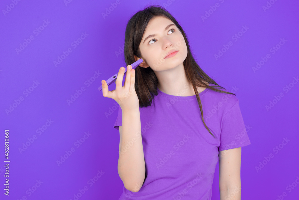Smiling young beautiful Caucasian woman wearing purple T-shirt over purple wall listening a voice message from her smartphone. Communication and technology concept. Smiling young beautiful Caucasian woman wearing purple T-shirt over purple wall listening a voice message from her smartphone. Communication and technology concept.