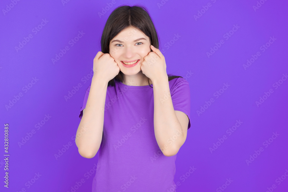 Happy young beautiful Caucasian girl wearing purple T-shirt over purple background keeps fists on cheeks smiles broadly and has positive expression being in good mood Happy young beautiful Caucasian girl wearing purple T-shirt over purple background keeps fists on cheeks smiles broadly and has positive expression being in good mood