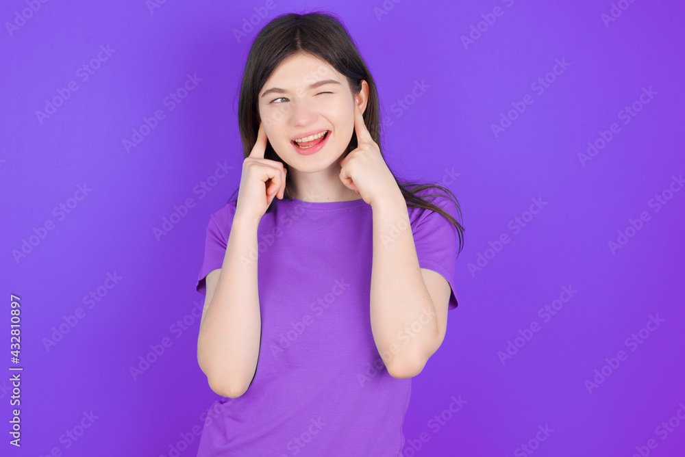 Happy young beautiful Caucasian girl wearing purple T-shirt over purple background ignores loud music and plugs ears with fingers asks to turn off sound