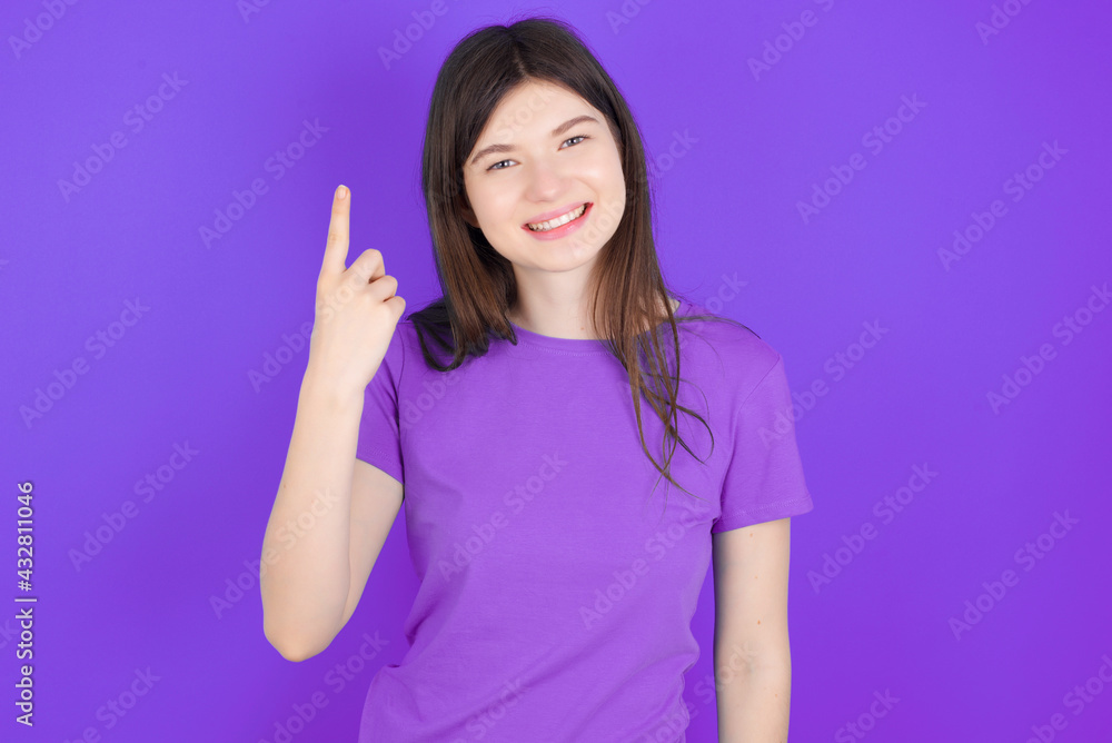 young beautiful Caucasian girl wearing purple T-shirt over purple background smiling and looking friendly, showing number one or first with hand forward, counting down young beautiful Caucasian girl wearing purple T-shirt over purple background smiling and looking friendly, showing number one or first with hand forward, counting down