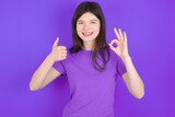 young beautiful Caucasian girl wearing purple T-shirt over purple background smiling and looking happy, carefree and positive, gesturing victory or peace with one hand