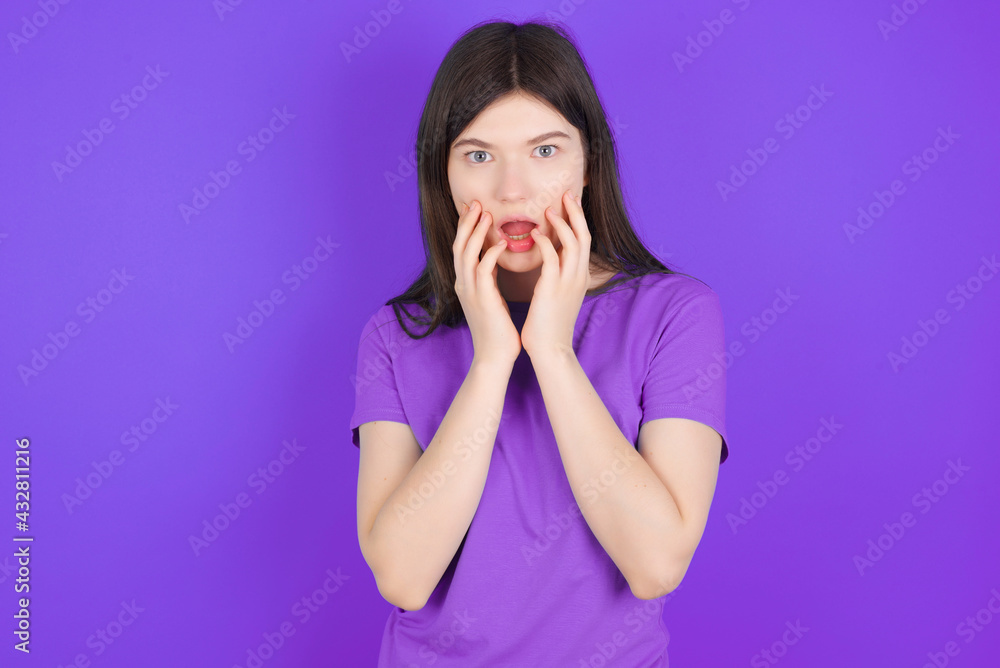 Speechless young beautiful Caucasian girl wearing purple T-shirt over purple background keeps hands near opened mouth reacts to shocking news stares wondered at camera Speechless young beautiful Caucasian girl wearing purple T-shirt over purple background keeps hands near opened mouth reacts to shocking news stares wondered at camera