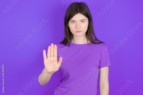 young beautiful Caucasian girl wearing purple T-shirt over purple background shows stop sign prohibition symbol keeps palm forward to camera with strict expression