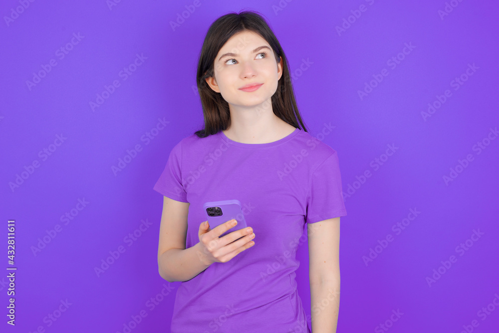 young beautiful Caucasian girl wearing purple T-shirt over purple background holds mobile phone uses high speed internet and social networks has online communication. Modern technologies concept young beautiful Caucasian girl wearing purple T-shirt over purple background holds mobile phone uses high speed internet and social networks has online communication. Modern technologies concept