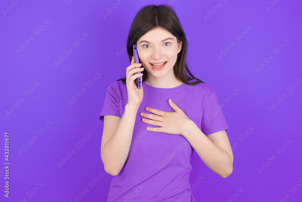 Smiling young beautiful Caucasian girl wearing purple T-shirt over purple background talks via cellphone, enjoys pleasant great conversation. People, technology, communication concept Smiling young beautiful Caucasian girl wearing purple T-shirt over purple background talks via cellphone, enjoys pleasant great conversation. People, technology, communication concept