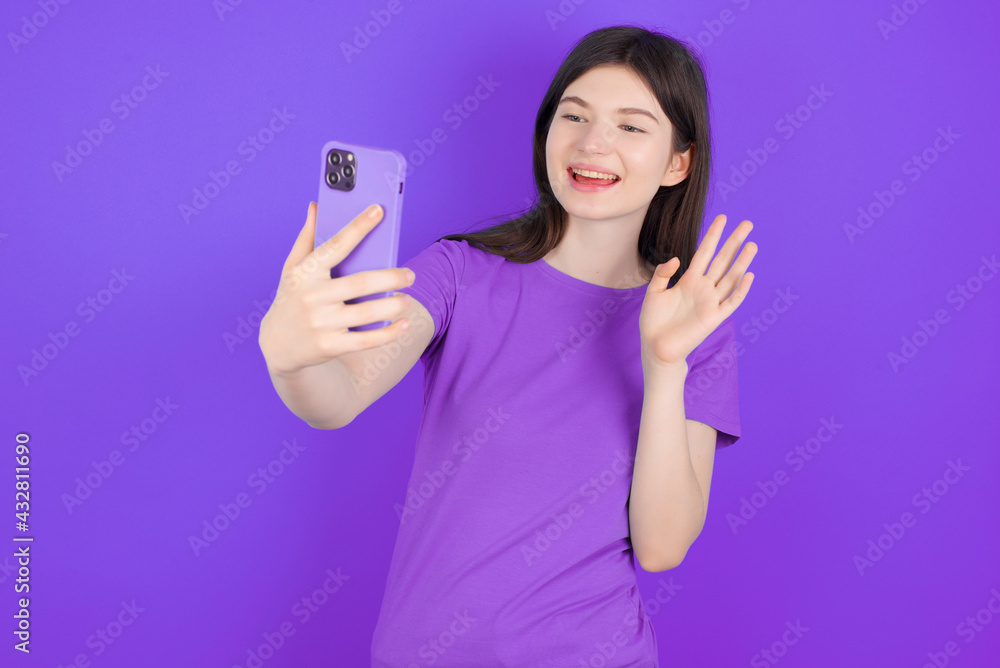 young beautiful Caucasian girl wearing purple T-shirt over purple background holds modern mobile phone and makes video call waves palm in hello gesture. People modern technology concept young beautiful Caucasian girl wearing purple T-shirt over purple background holds modern mobile phone and makes video call waves palm in hello gesture. People modern technology concept