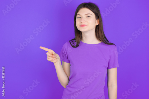 young beautiful Caucasian girl wearing purple T-shirt over purple background points to side on blank space demonstrates advertisement. People and promotion concept