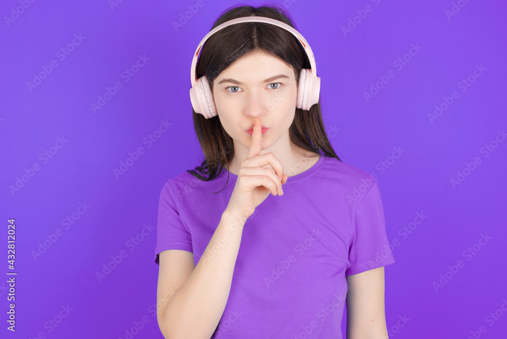 young beautiful Caucasian girl wearing purple T-shirt over purple background making hush gesture with finger on her lips wearing wireless headphones. Be quiet. young beautiful Caucasian girl wearing purple T-shirt over purple background making hush gesture with finger on her lips wearing wireless headphones. Be quiet.