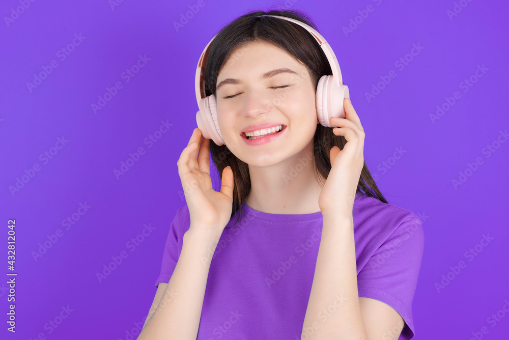 young beautiful Caucasian girl wearing purple T-shirt over purple background with headphones on her head, listens to music, enjoying favourite song with closed eyes, holding hands on headset. young beautiful Caucasian girl wearing purple T-shirt over purple background with headphones on her head, listens to music, enjoying favourite song with closed eyes, holding hands on headset.