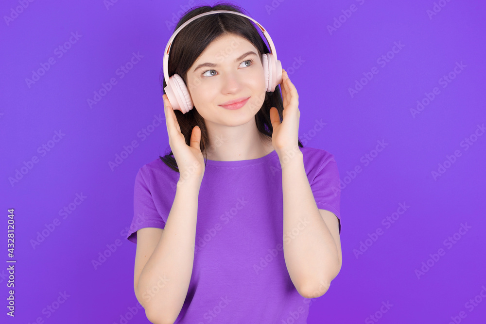 young beautiful Caucasian girl wearing purple T-shirt over purple background wears stereo headphones listens music concentrated aside. People hobby lifestyle concept young beautiful Caucasian girl wearing purple T-shirt over purple background wears stereo headphones listens music concentrated aside. People hobby lifestyle concept
