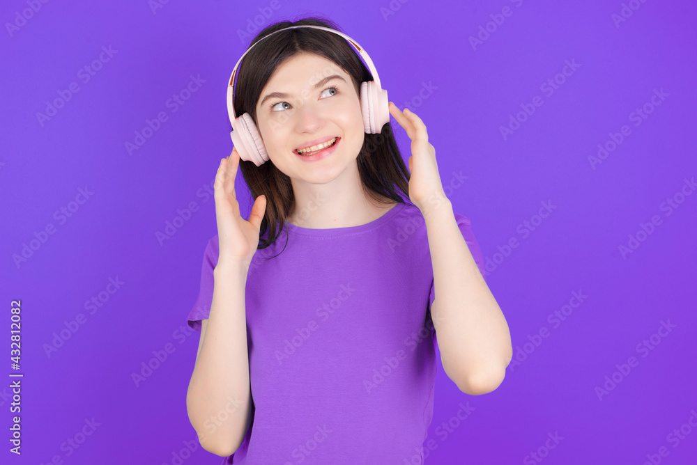 Joyful young beautiful Caucasian girl wearing purple T-shirt over purple background sings song keeps hand near mouth as if microphone listens favorite playlist via headphones Joyful young beautiful Caucasian girl wearing purple T-shirt over purple background sings song keeps hand near mouth as if microphone listens favorite playlist via headphones