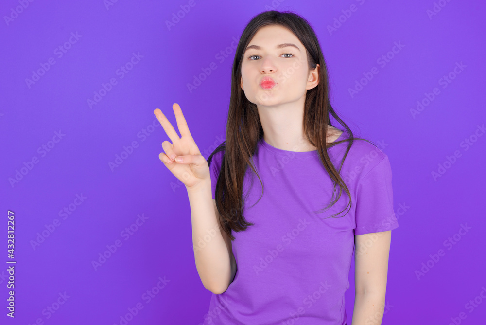 young beautiful Caucasian girl wearing purple T-shirt over purple background makes peace gesture keeps lips folded shows v sign. Body language concept young beautiful Caucasian girl wearing purple T-shirt over purple background makes peace gesture keeps lips folded shows v sign. Body language concept