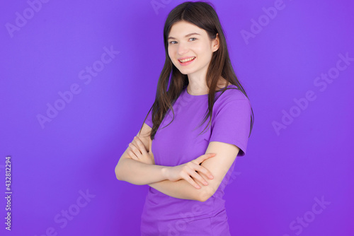 Portrait of young beautiful Caucasian girl wearing purple T-shirt over purple background standing with folded arms and smiling