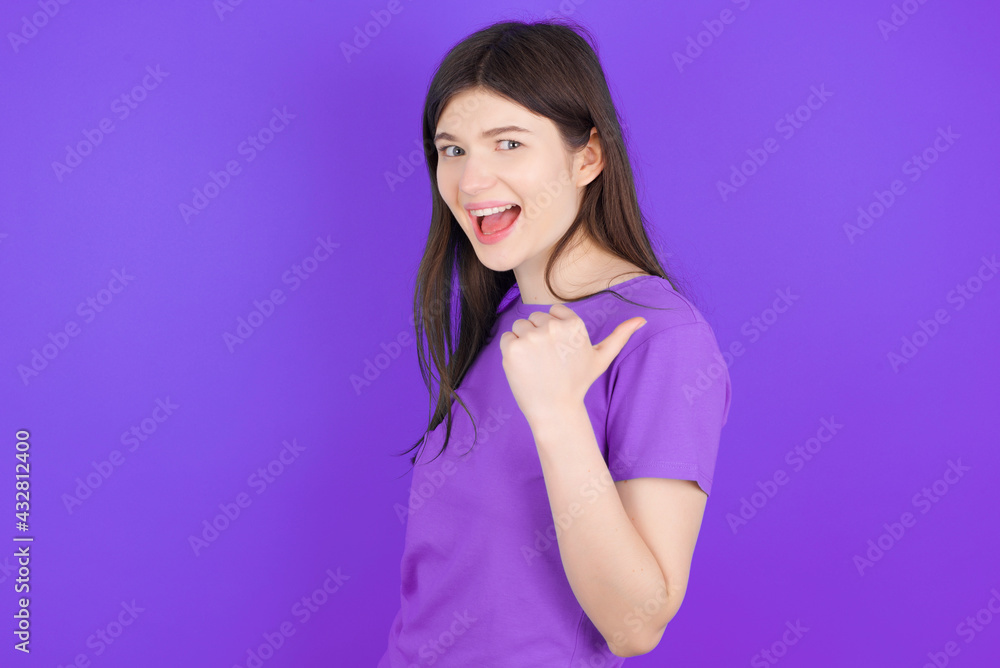 Impressed young beautiful Caucasian girl wearing purple T-shirt over purple background point back empty space Impressed young beautiful Caucasian girl wearing purple T-shirt over purple background point back empty space