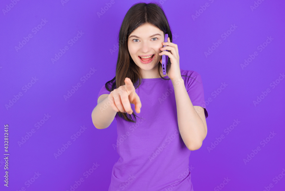 Positive young beautiful Caucasian girl wearing purple T-shirt over purple background indicates directly at camera has telephone conversation smiles broadly enjoys talking long hours. You join me Positive young beautiful Caucasian girl wearing purple T-shirt over purple background indicates directly at camera has telephone conversation smiles broadly enjoys talking long hours. You join me
