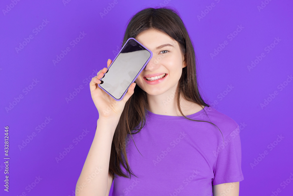 young beautiful Caucasian girl wearing purple T-shirt over purple background holding modern smartphone covering one eye while smiling young beautiful Caucasian girl wearing purple T-shirt over purple background holding modern smartphone covering one eye while smiling