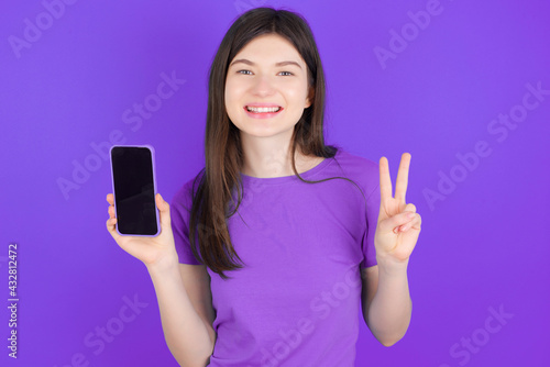 young beautiful Caucasian girl wearing purple T-shirt over purple background holding modern device showing v-sign
