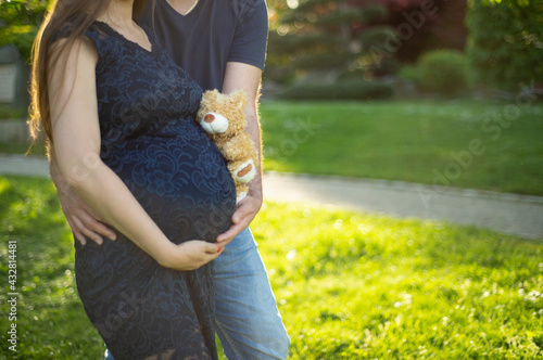 Ein werdender Vater umarmt vorsichtig seine schwangere Frau, die einen kleinen Teddybär an Ihren Bauch drückt