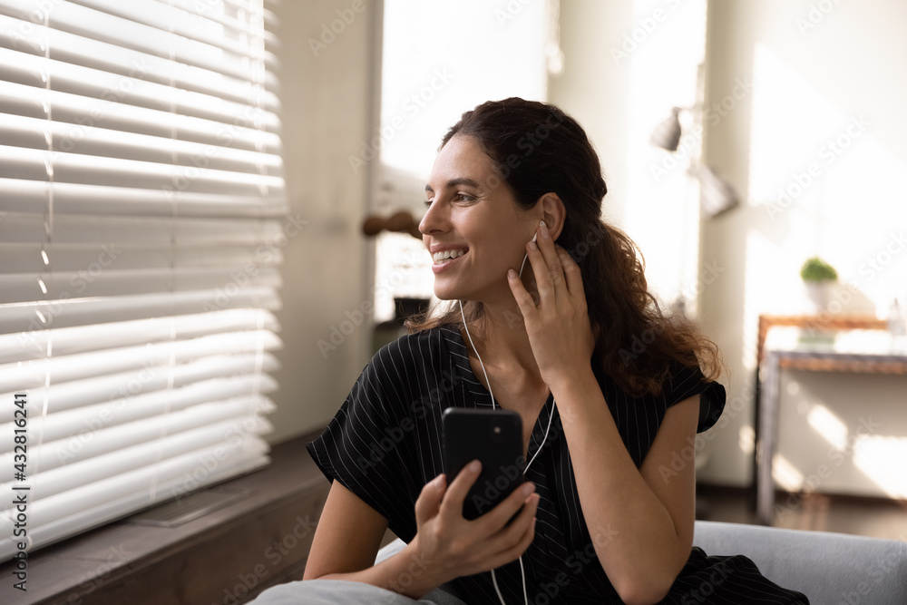 Lost in pleasant dreams. Smiling young woman in headset relax on sofa with smartphone look at window think distracted from device screen. Happy latin female hold cell listen to good music in earphones Lost in pleasant dreams. Smiling young woman in headset relax on sofa with smartphone look at window think distracted from device screen. Happy latin female hold cell listen to good music in earphones