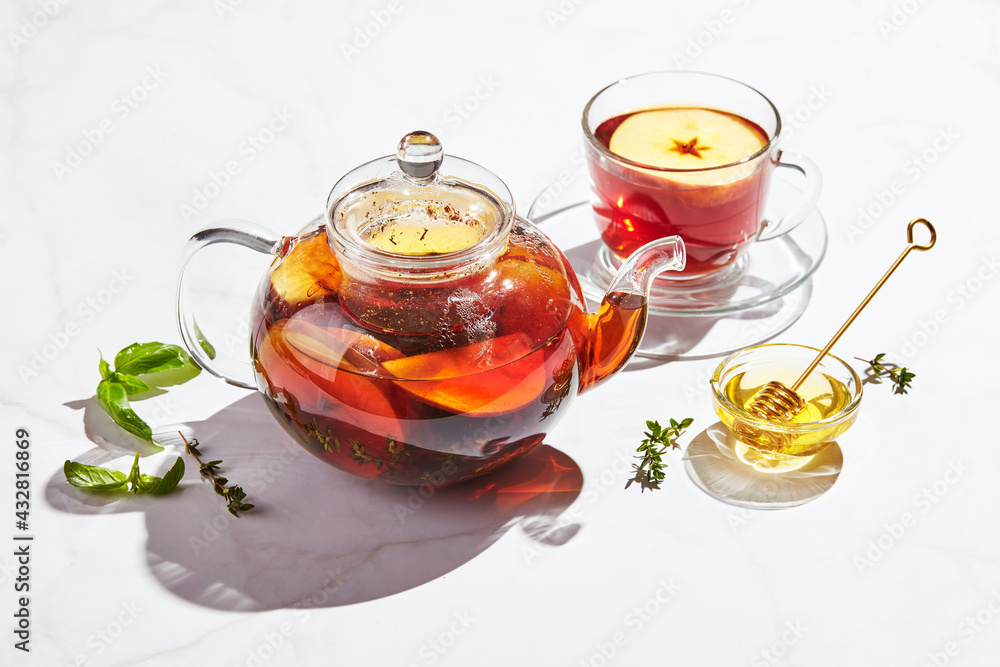 Fruit tea with apples and thyme and honey in glass teapot and cup on white background with hard shadows Fruit tea with apples and thyme and honey in glass teapot and cup on white background with hard shadows