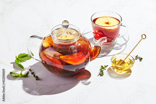 Fruit tea with apples and thyme and honey in glass teapot and cup on white background with hard shadows