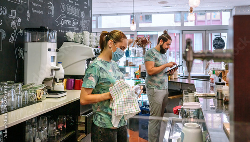 Waitress with mask cleaning glasses in a coffee shop while her coworker works with a tablet
