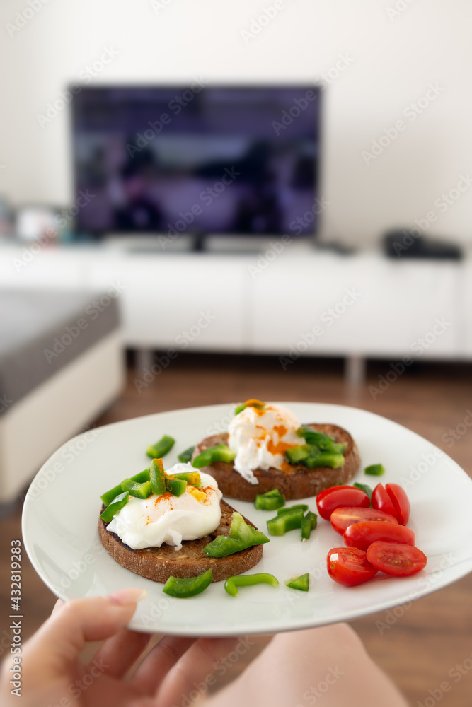 Close up of female hand holding a white plate with whole grain toasted bread and pouched eggs with cherry tomatoes and green peppers, interiour of living room, lifestyle concept Close up of female hand holding a white plate with whole grain toasted bread and pouched eggs with cherry tomatoes and green peppers, interiour of living room, lifestyle concept