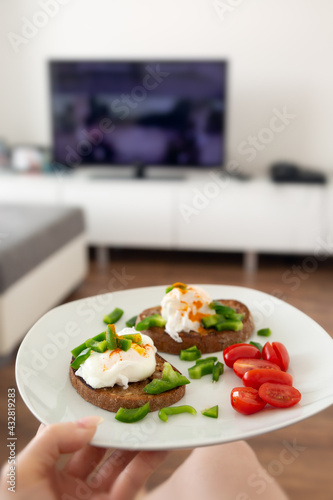 Close up of female hand holding a white plate with whole grain toasted bread and pouched eggs with cherry tomatoes and green peppers, interiour of living room, lifestyle concept